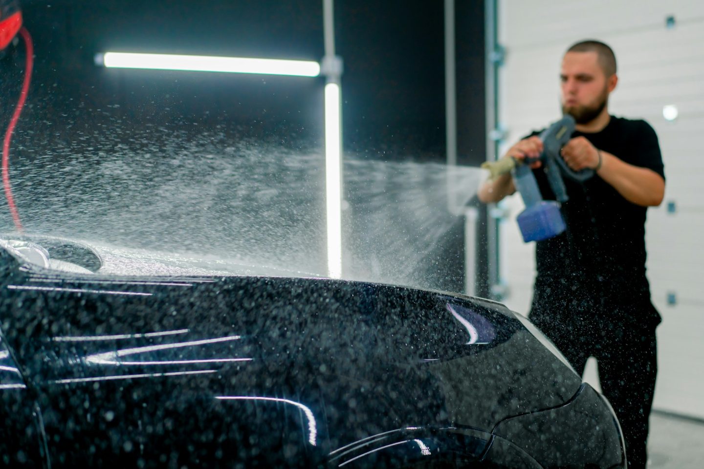 A male car wash employee applies car wash foam to a luxury black car using spray gun in car wash box