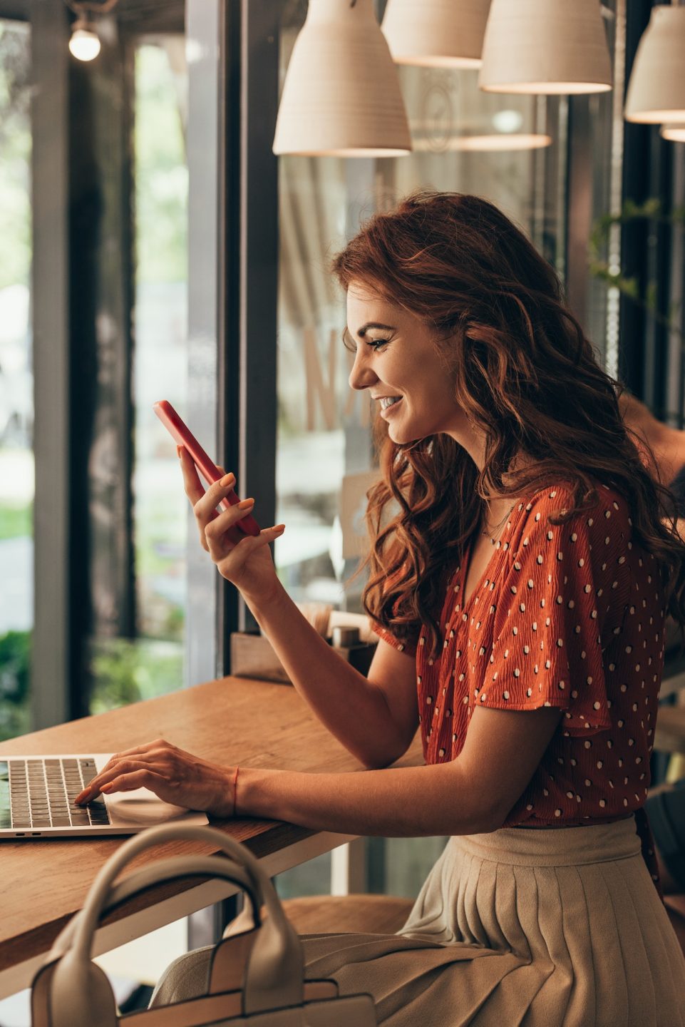 side view of cheerful young woman using smartphone at table with laptop in coffee shop